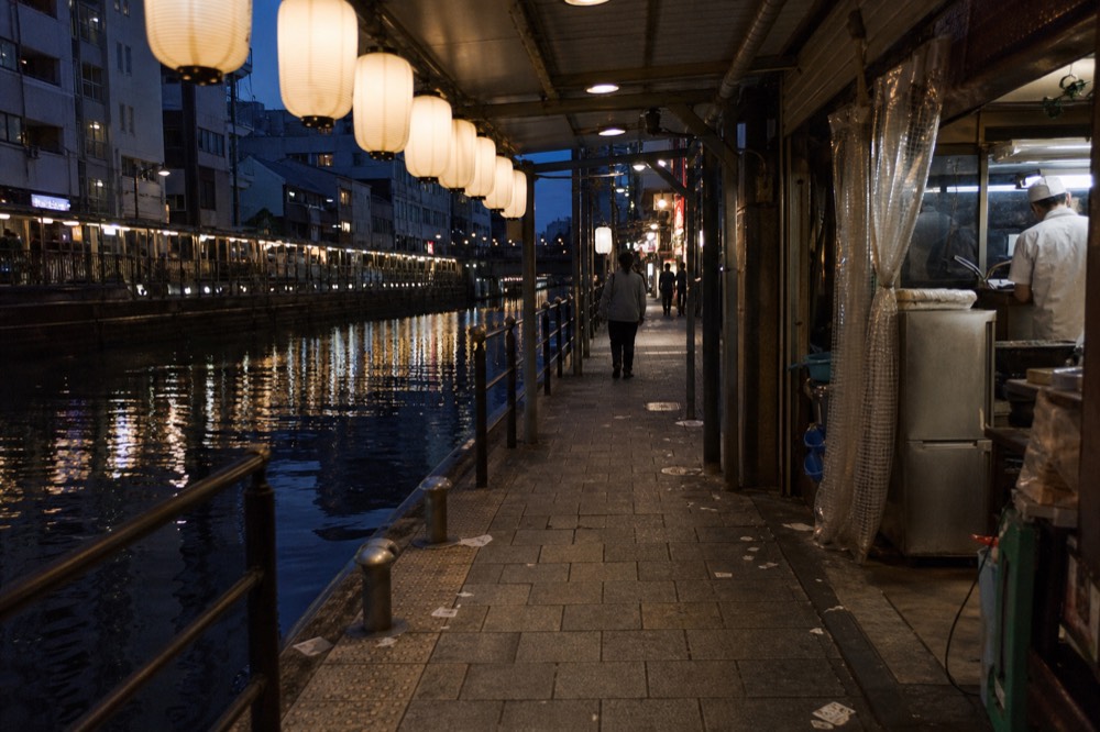 A covered shopping street in the Dotonbori area with illuminated signs along the canal