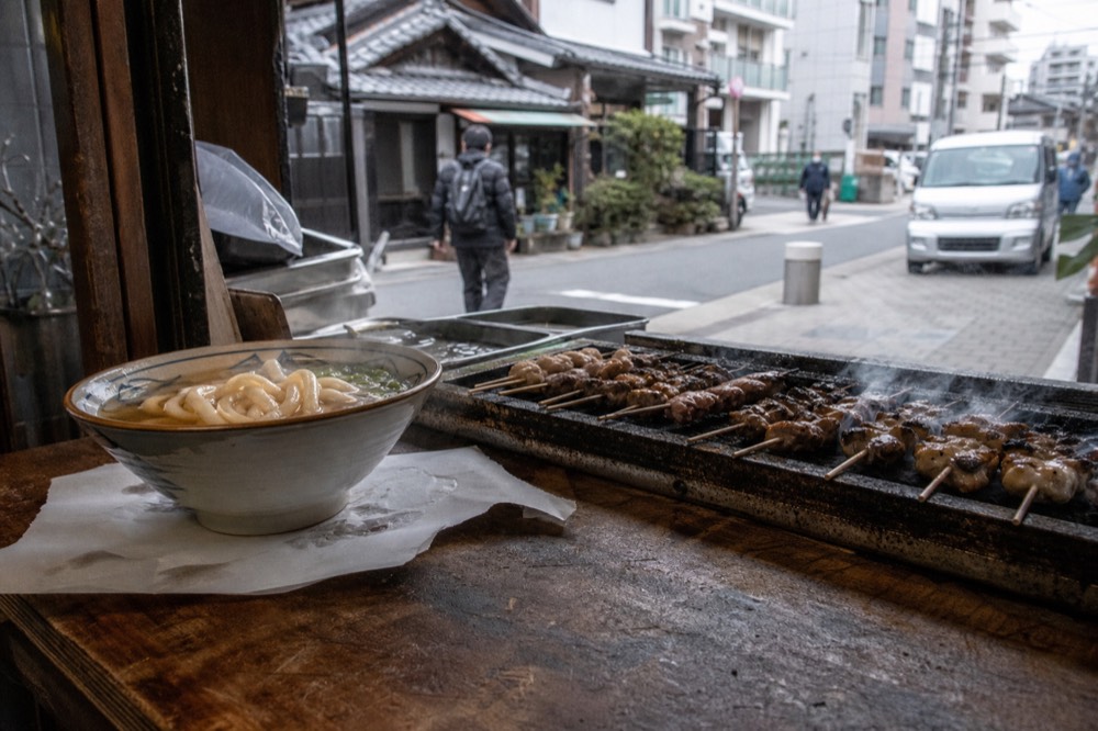 Bowl of noodles and grilled skewers on a wooden counter at a food stall